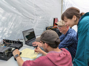 Two men and a woman work on a ham radio and laptop in a tent