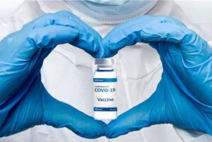 Hands in blue medical gloves, shaped as a heart, holding a vial of COVID vaccine in the center
